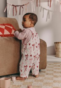 Bela kombinezon z rdečimi in roza vzorci pentelj, nošen prek roza majice z dolgimi rokavi. Otrok stoji ob rjavi kavču, bos na teksturnem preprogi.