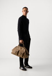 Brown leather duffel bag with two front pockets, zipper closure, and sturdy handles. Worn with black joggers and black shoes with white stripes.
