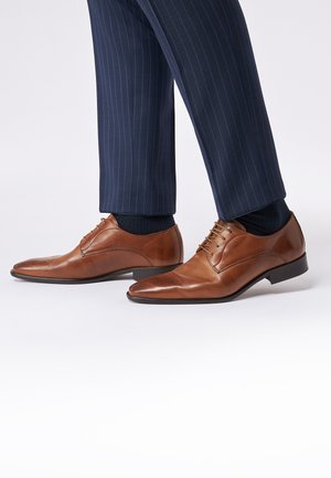 Brown leather dress shoes with a polished finish, pointed toe, and perforated detailing, paired with navy pinstripe trousers and dark socks.