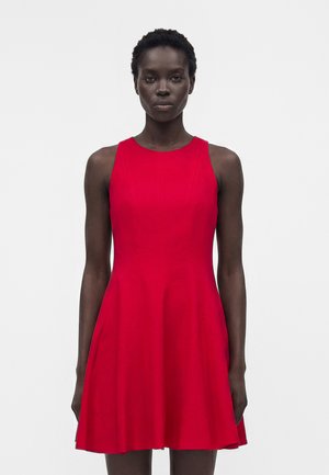 Kvinde med kort krøllet hår iført en ærmeløs, knælængde rød kjole, stående mod en ensfarvet lys baggrund.