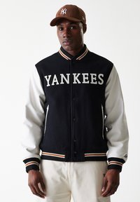 Svart og hvit ulljakke med "Yankees"-tekst, ribbet krage og mansjetter, knappelukking og sidelommer. Brun kordfløyels-cap.