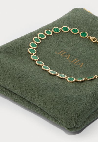 Pulsera dorada con piedras ovaladas de rayas verdes, reposando sobre un pouch de gamuza verde con "JIAJIA" embellesado en dorado.
