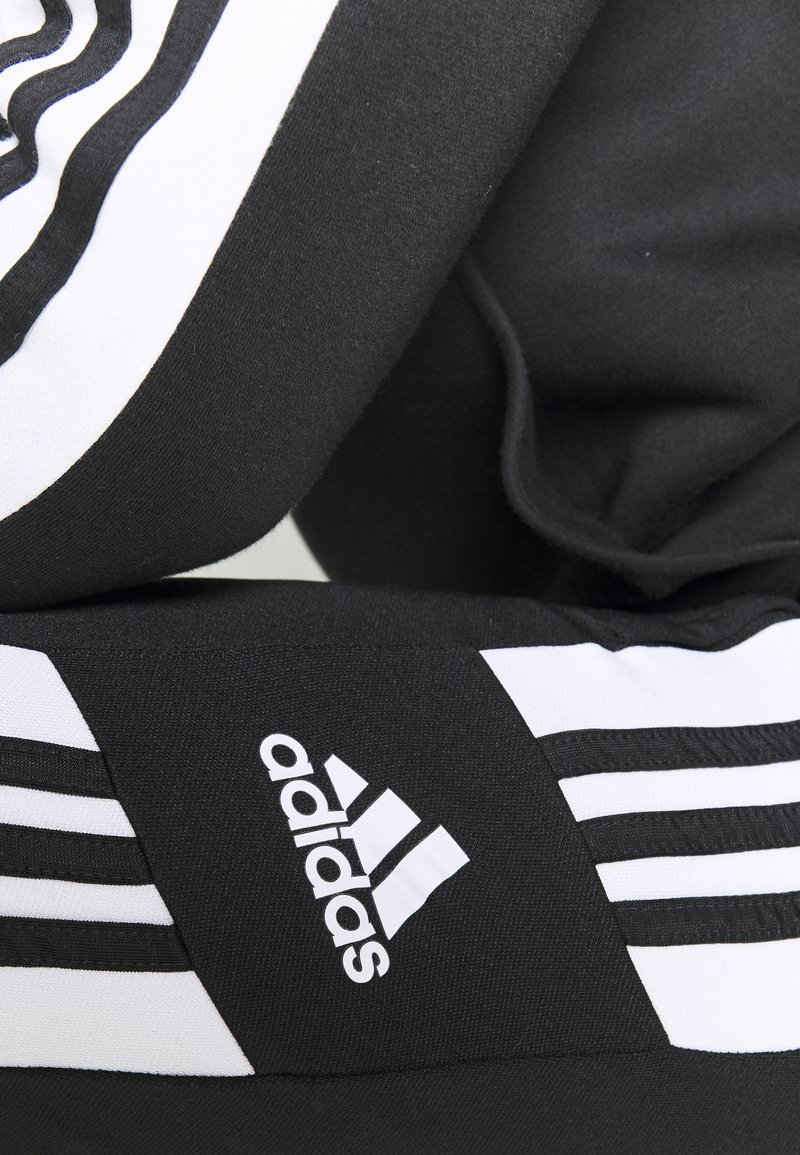 Pantalons de sport noirs avec des accents blancs et un design à trois bandes, fabriqués en tissu texturé. Affiche un logo Adidas sur le côté.