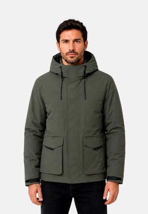 Homme portant une veste d'hiver verte à capuche avec des poches avant et des cordons noirs, debout devant un fond clair uni.