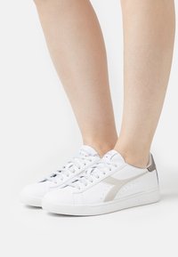 Baskets blanches à lacets avec une texture lisse ; présentent une accent beige et des perforations sur le bout. Semelle en caoutchouc, design minimaliste.