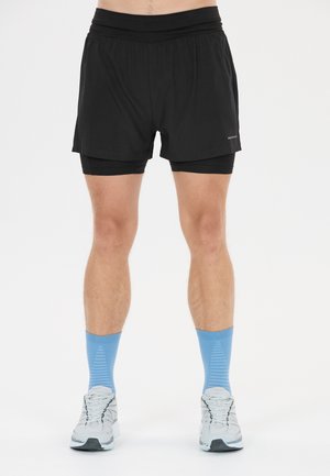 Mann trägt schwarze, geschichtete Laufshorts, hellblaue Sportsocken und weiße Laufschuhe und steht vor einem schlichten Hintergrund.