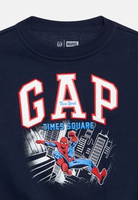 Μπλε σκούρο φούτερ με κόκκινα και λευκά γράμματα "GAP", με γραφικό του Spider-Man μπροστά σε φόντο ορίζοντα πόλης. Μαλακό βαμβακερό υλικό.