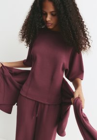 Maroonský sportovní set obsahující volnější top s spadlými rameny a širokým výstřihem, doplněný o úzké kalhoty s nastavitelným stahováním.
