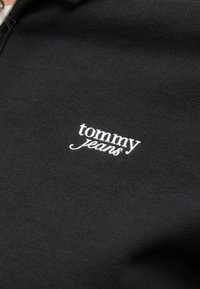 Tela negra con el logo "tommy jeans" bordado en blanco en letras minúsculas centrado en el material.