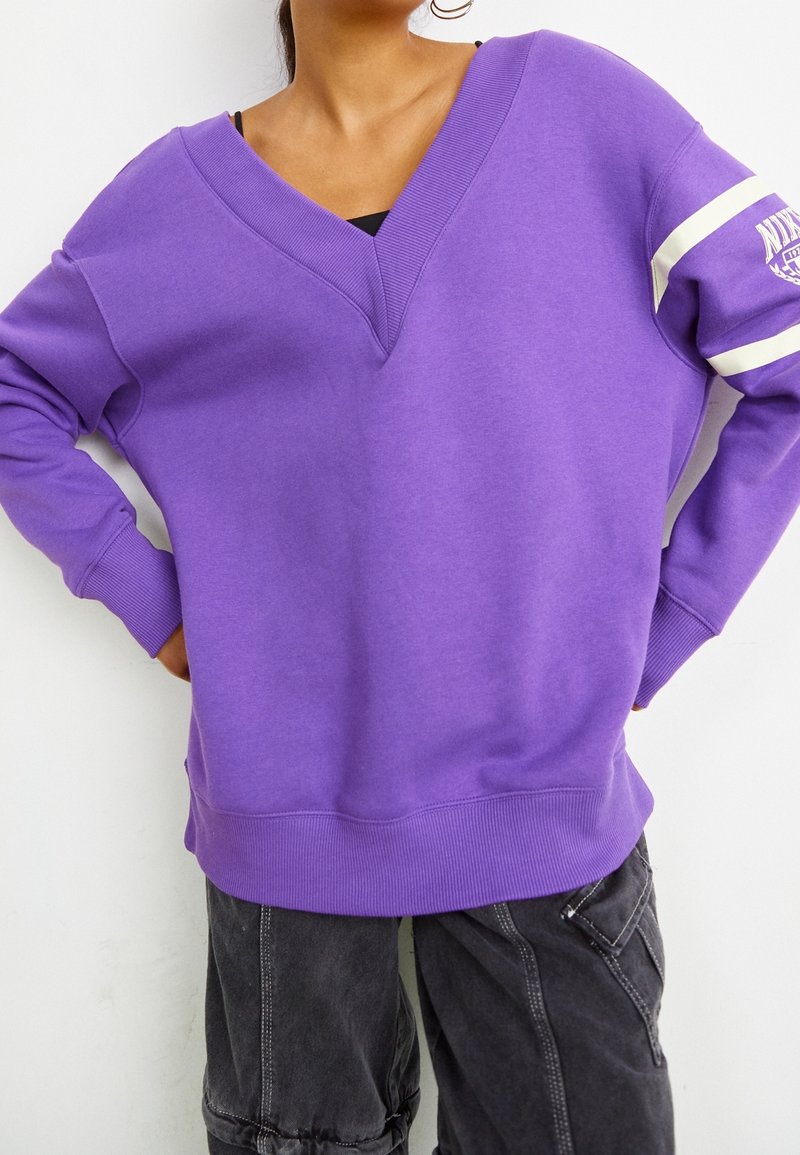 Femme portant un sweat-shirt violet oversize à col en V avec des rayures blanches et un logo sur la manche, ainsi qu’un pantalon cargo noir, debout les mains sur les hanches.