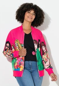 Pink bomberjakke med livlige, tegneserie-stil kvindelige illustrationer på ærmerne. Sort v-hals top under, båret med blå jeans.