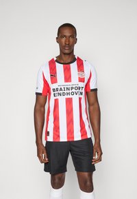 Rød- og hvidstribet fodboldtrøje med et chevronmønster, sorte detaljer og sponsorlogo; parret med sorte shorts og hvide sokker.