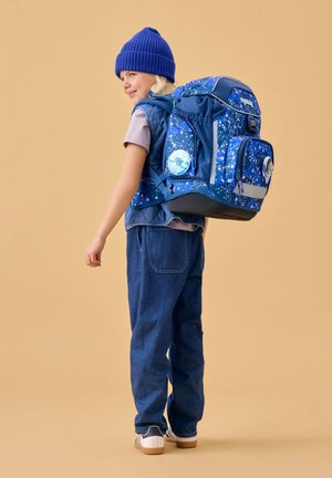 Child in blue beanie, denim pants, and vest carries large blue backpack with patterned design and bird-themed patches.