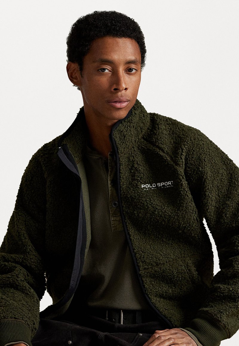 Olivgrüne Fleecejacke mit strukturierten Oberflächen und Reißverschluss, ausgestattet mit einem schwarzen Akzentrand und dem "POLO SPORT"-Logo auf der Brust.