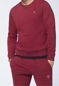 Sudadera burdeos con puños y dobladillo acanalados, que presenta un pequeño detalle bordado en el pecho. Combinada con pantalones jogger a juego.