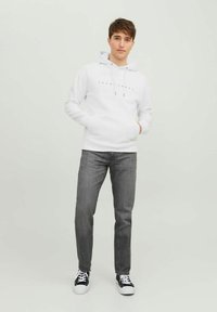 Sweat à capuche blanc avec cordons et logo imprimé, associé à un jean gris et des baskets noires. Le modèle se tient contre un fond clair.