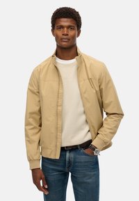Beige letvægtsjakke med ståkrave, frontlynlås og elastiske manchetter. Båret over en lys sweater og parret med blå jeans.