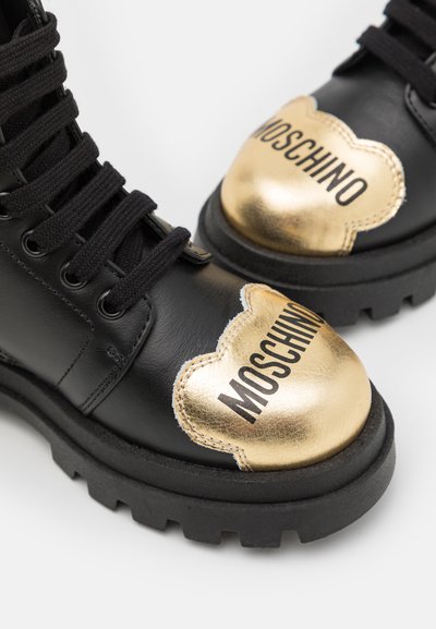 Botas de cuero negro con una suela gruesa negra, que presentan una puntera curva dorada y "MOSCHINO" impreso en negro sobre la superficie dorada.