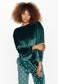 Top verde scuro in velluto con maniche lunghe e scollatura ampia, abbinato a pantaloni a pois nei toni di verde coordinati. Tessuto morbido e liscio.