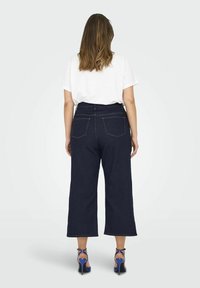 Mörkblå, vida denimjeans med hög midja, bakfickor och synlig gul sömnad, kombinerat med en vit t-shirt och blå klacksandaler.