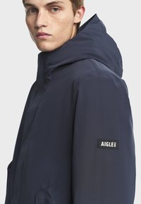 Mladý muž ve tmavě modré mikině s kapucí, na které je nášivka s logem AIGLE 1853 na rukávu, obrácený mírně doleva před jednobarevným pozadím.