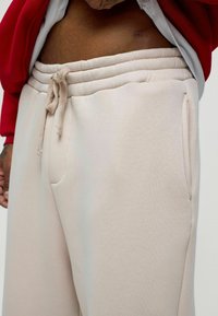 Beige sweatpants met elastische tailleband en trekkoord, met zijzakken en een zachte, gestructureerde stof. Rood sweatshirt zichtbaar erboven.