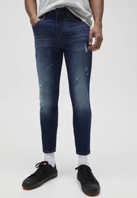 Vaqueros azul denim de corte ajustado con ligera decoloración y detalles desgastados, cuentan con cinco bolsillos y cierre clásico de botón.