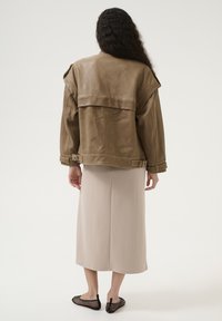Jachetă din piele maro cu guler supradimensionat, două accente cu cataramă, asortată cu o fustă bej și pantofi slip-on din plasă negri.
