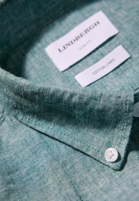 Camicia verde in cotone e lino con vestibilità slim; presenta un colletto a bottone e bottoni bianchi. Le etichette indicano il marchio e la composizione del materiale.