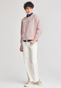 Felpa rosa chiaro con la scritta "Polo Club" stampata in rosa scuro, vestibilità ampia, polsini a coste, indossata sopra una camicia di jeans scuri, abbinata a pantaloni bianchi e sneakers beige.