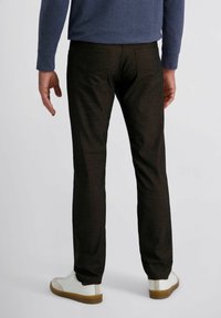 Persona con pantaloni neri slim, sneakers bianche con suole marroni e una camicia a maniche lunghe blu, in piedi contro uno sfondo neutro.