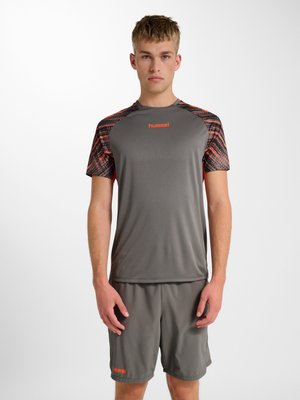 Giovane uomo con indosso abbigliamento sportivo grigio con accenti arancioni, in piedi di fronte a uno sfondo bianco semplice.
