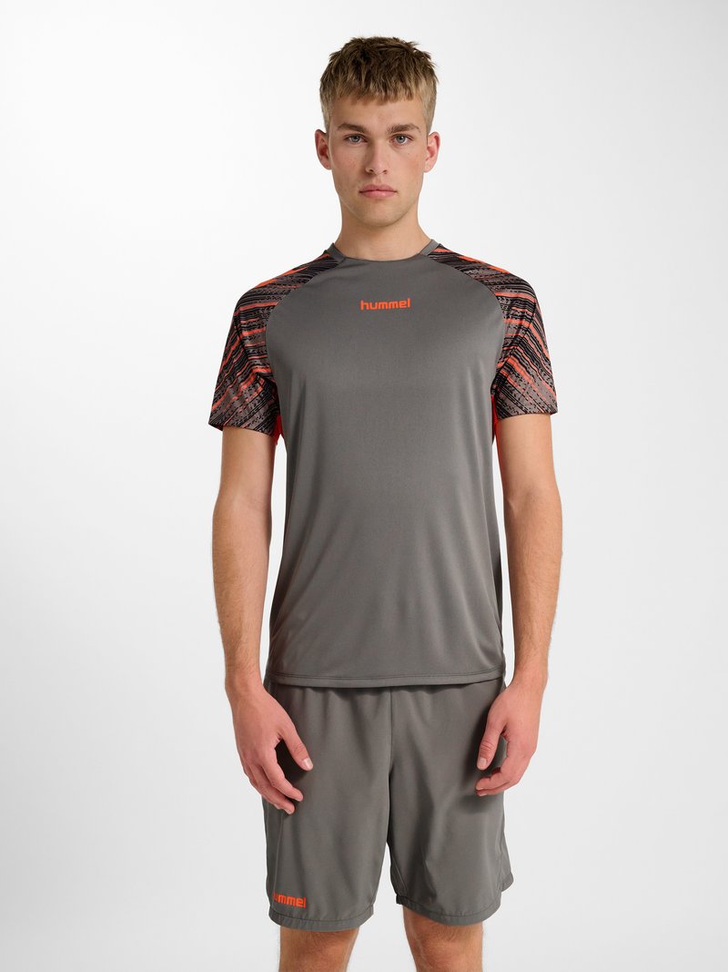 Jonge man in grijze sportkleding met oranje accenten, recht vooruit kijkend tegen een effen witte achtergrond.