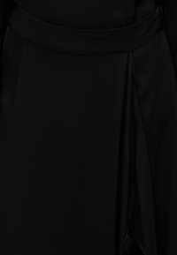 Μαύρο φόρεμα με απαλό, ρέον ύφασμα, ζώνη στη μέση και σκίσιμο στο πλάι. Η υφή φαίνεται μαλακή και ελαφριά.