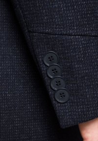 Ciemnoniebieska, teksturowana tkanina rękawa marynarki z czterema czarnymi guzikami z dwiema dziurkami każdy, obok szczegółu szwa małej kieszonki.