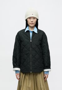 Chaqueta negra acolchada con cremallera plateada, usada sobre una camisa azul claro. Gorro de punto crema en la cabeza. Falda con diseño plisado.