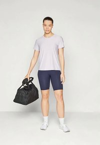 Hele lilla T-särk on kombineeritud tumepruunide jalgrattashortsidega. Mudel hoiab käes musta spordikoti, kandes valgeid spordijalgu ja pahkluu sokke.