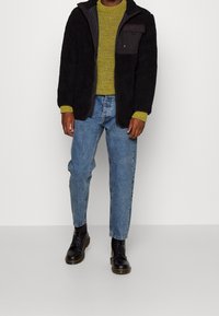 Černá fleecová bunda, olivově zelený pletený svetr, světle modré džíny a černé šněrovací boty. Mezi rysy patří zip a náprsní kapsa.