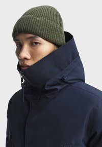 Marineblauwe jas met een hoge kraag, voorzien van een rits en een verstelbaar koord. Groene geribbelde beanie bovenop, close-up van het kraaggebied.