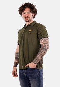 Polo color verde oliva con cuello, mangas cortas y un acento de logo. Textura suave con ribetes contrastantes en el cuello y las mangas.