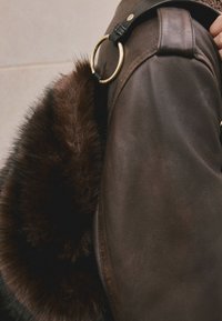 Épaule de veste en cuir marron avec sangle noire et détail d'anneau doré, à côté d'une texture de fourrure brun foncé.
