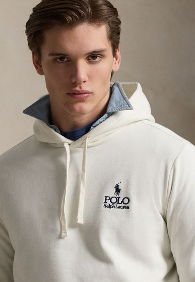 Νεαρός άνδρας που φοράει μια κρεμ φούτερ Polo Ralph Lauren πάνω από ένα μπλε πουκάμισο με γιακά, κοιτάζοντας κατευθείαν την κάμερα με ουδέτερη έκφραση.