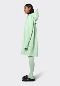 Mintgroene waterdichte jas met een capuchon, ontspannen pasvorm, lange mouwen, gecombineerd met bijpassende leggings en zwarte schoeisel. Gladde textuur, minimalistisch ontwerp.