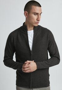 Giacca a zip grigio scuro con design a coste testurizzato, colletto alto e silhouette aderente. Presenta un interno liscio e dettagli a coste.