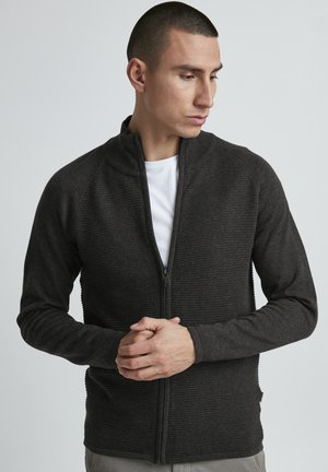 Mørkegrå zip-up jakke med en teksturert ribbestrikket design, høy krage og tettsittende silhuett. Har et glatt interiør og ribbestrikkede detaljer.