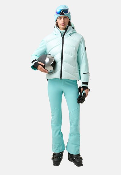 Eine hellblaue Pufferjacke über passenden Skihosen, mit einem silbernen Helm in der Hand und schwarzen Handschuhen, vor einem neutralen Hintergrund.