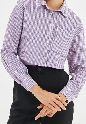 Persoon die een paars geruit overhemd met knopen draagt, ingestopt in een zwarte broek met hoge taille, met ringen aan de vingers en handen gekruist ter hoogte van de taille.