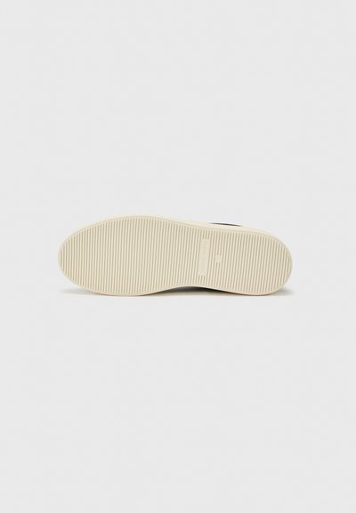 Mustan lenkkarin pohja, jossa on teksturoitu pinta ja pystysuorat urat, ja kontrastoiva vaaleanbeige kumipohja. Minimalistinen muotoilu logotunnuksella.