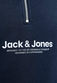 Marin zip-up sweatshirt med en texturerad tyg. Har en silverdragkedja och vittryckt text: "Jack & Jones" samt varumärkesdetaljer.