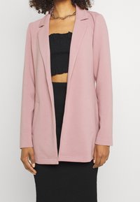 Różowa marynarka z długim rękawem i klapami z wycięciem, wykonana z teksturowanej tkaniny, wyposażona w boczne kieszenie i luźny fason, noszona na czarnej bluzce.